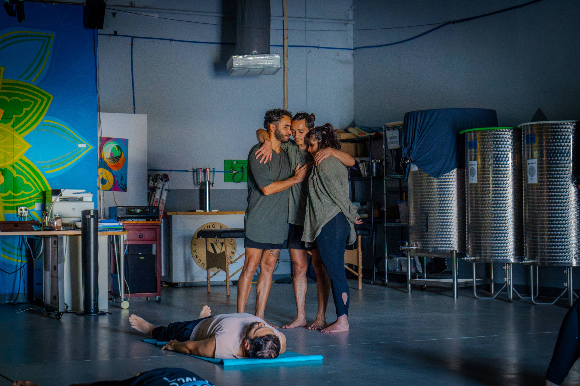 Three practitioners holding each other in a quiet embrace while another rests on a mat below